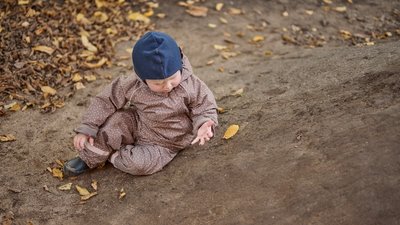 Ein Kleinkind in brauner, gepunkteter Outdoor-Kleidung und blauer Mütze sitzt auf einem erdigen Boden im Freien und betrachtet ein Herbstblatt in seiner Hand; rundherum liegen weitere gelbe und braune Blätter.