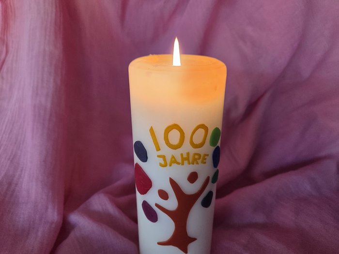Brennende Jubiläumskerze mit der Aufschrift ‚100 Jahre und farbigem Baum-Motiv, vor weichem rosafarbenem Hintergrund.