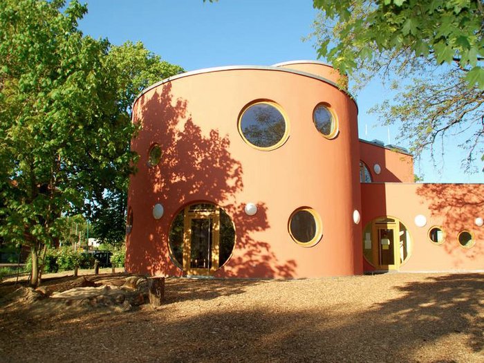 Das Gebäude „Schildow“ mit runder, terrakottafarbener Fassade und mehreren kreisförmigen Fenstern steht freistehend in einer grünen Umgebung mit Bäumen; vor dem Eingang liegt ein mit Holzspänen bedeckter Außenbereich bei sonnigem Wetter.