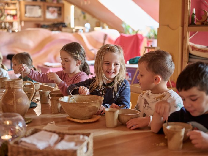 Mehrere Kinder sitzen an einem langen Holztisch und rühren mit Löffeln in Schalen und Bechern; sie sind konzentriert und im Austausch miteinander, umgeben von Keramikgeschirr und Materialien in einem warm gestalteten Innenraum.