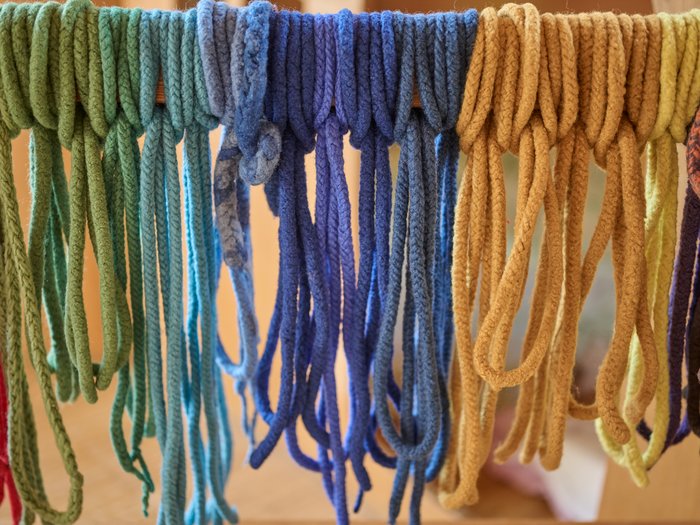 Mehrere dicke, weiche Woll- oder Garnstränge hängen in Schlaufen über einer Stange; sie sind nach Farben sortiert und verlaufen von Grün- und Türkistönen über Blau bis zu Gelb- und Ockertönen vor einem unscharfen Hintergrund.