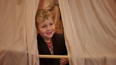 Ein lächelndes Kind blickt durch einen Spalt zwischen zwei hellen Stoffbahnen; es hält eine Holzstange in den Händen und steht in einem warm beleuchteten Innenraum.