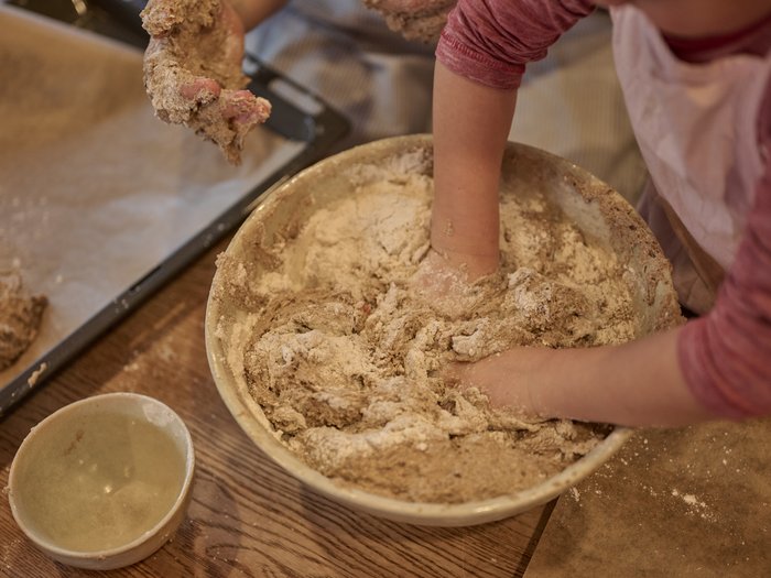 Kinderhände kneten eine mehlige Teigmasse in einer großen Schüssel; daneben stehen eine kleine Keramikschale und ein Backblech, alles auf einem Holztisch in einem Innenraum.