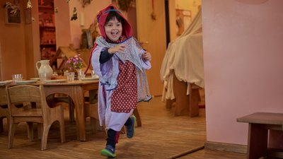 Ein lachendes Kind in bunter Verkleidung mit Kapuze läuft spielend durch einen warm beleuchteten Raum mit Holzboden; im Hintergrund stehen ein gedeckter Holztisch mit Geschirr sowie weitere Möbel und hängende Naturdekorationen.